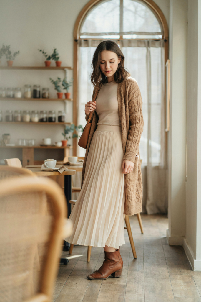 a girl in a a cream maxi skirt, fitted beige long-sleeve top, long camel knitted cardigan, brown ankle boots.