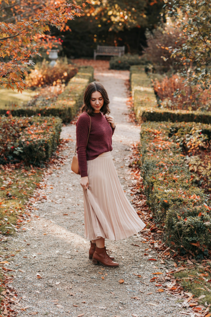 a girl in
a flowing pleated beige maxi skirt, fitted burgundy knit sweater, brown ankle boots, small shoulder bag, autumn garden with warm leaves, soft golden sunlight, elegant cozy styling.