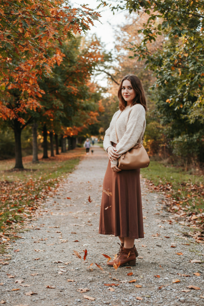 A girl in chocolate brown ankle-length maxi skirt paired with a soft cream oversized sweater, brown ankle boots, tan leather handbag.
