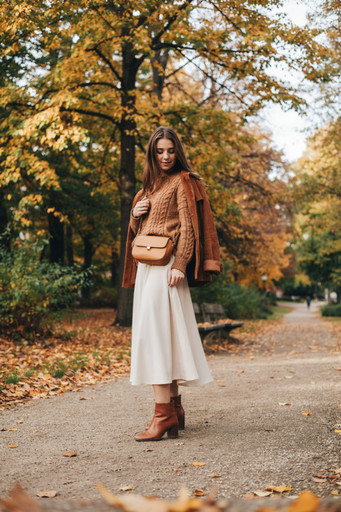 a girl in modest ivory ankle-length maxi skirt paired with a camel oversized sweater, brown ankle boots, tan crossbody bag, lightweight brown jacket, autumn park background with golden leaves,