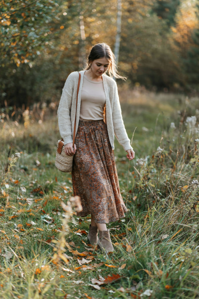 a girl with printed ankle-length maxi skirt in earthy brown and rust tones, cream knit cardigan, simple fitted top, suede ankle boots, woven handbag, autumn meadow background,
