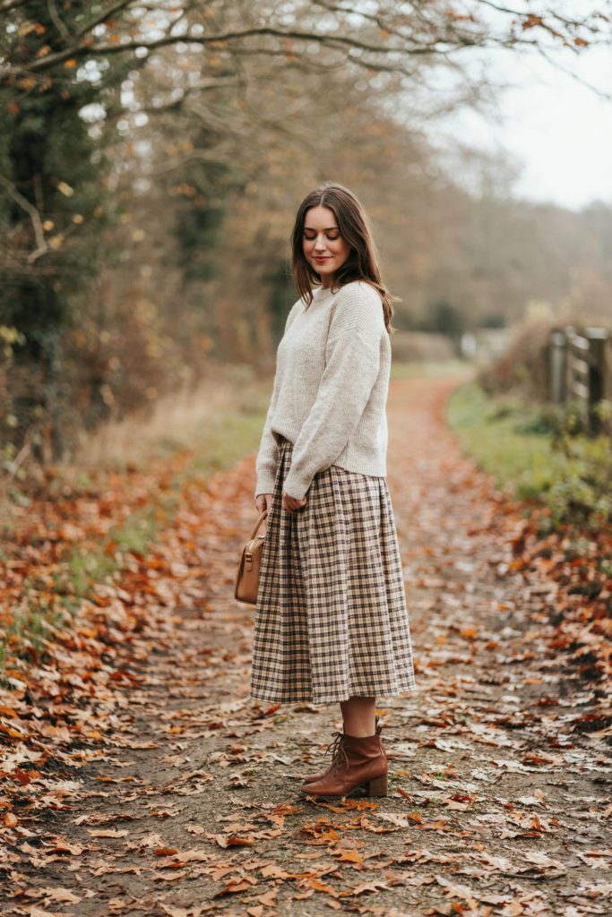A girl with ankle-length maxi skirt in brown, beige and cream, simple ivory knit sweater, brown ankle boots, small leather handbag