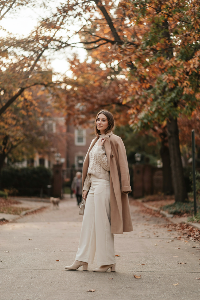 Woman wearing a cream knit sweater, ivory wide-leg trousers, long beige coat, cream ankle boots and neutral handbag, soft autumn background, elegant cozy feminine fashion, warm natural light.
