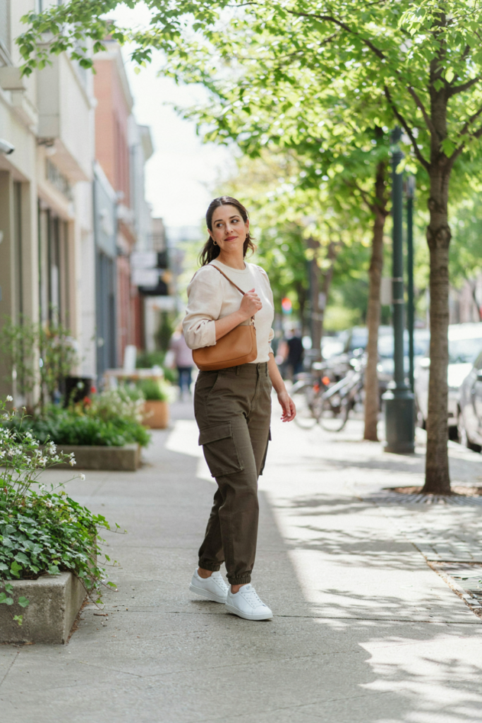 :
Curvy midsize woman wearing high-waisted olive cargo pants, soft cream fitted blouse with subtle puff sleeves, white sneakers, tan crossbody bag, and small gold earrings, modern spring city street with greenery