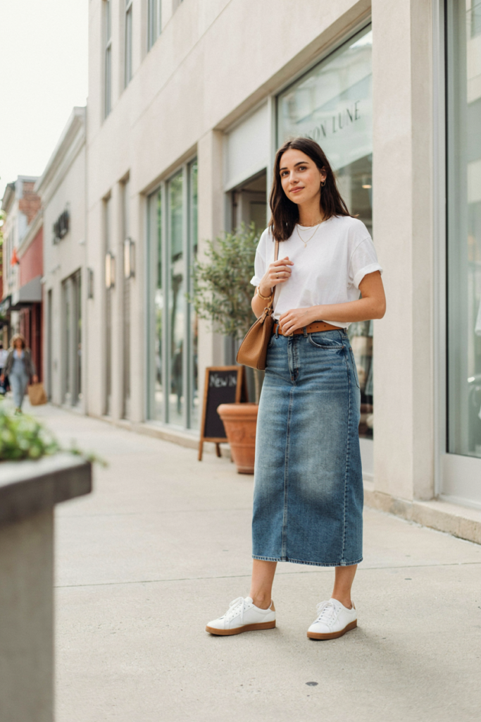 woman wearing a long denim midi skirt with a tucked-in soft white relaxed t-shirt, brown leather belt, white sneakers, small shoulder bag, subtle gold jewelry