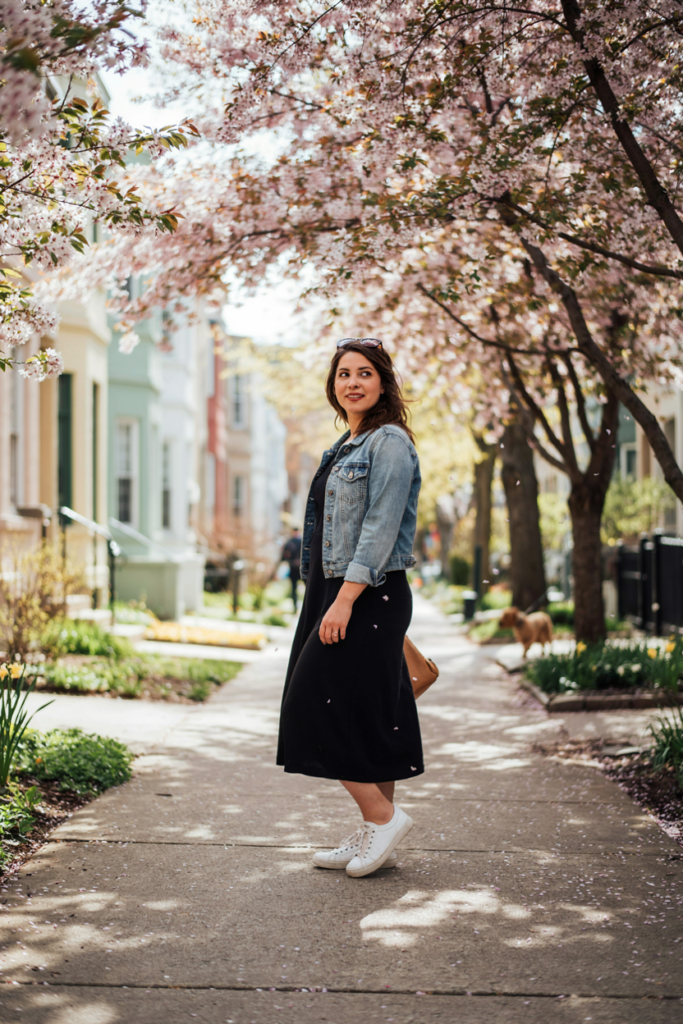 oman wearing a simple black midi dress, cropped light-wash denim jacket, white sneakers, small tan handbag, and sunglasses