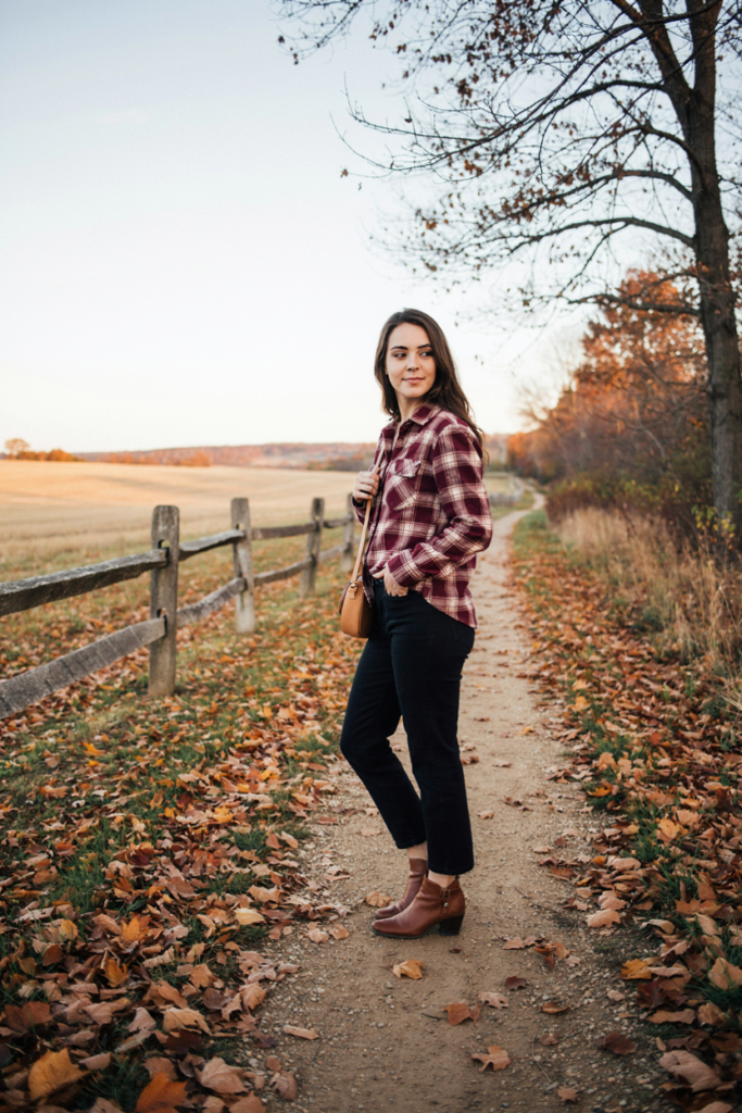 Woman wearing a burgundy and cream plaid shirt, black straight-leg jeans, brown ankle boots and crossbody bag, rustic autumn background with leaves.